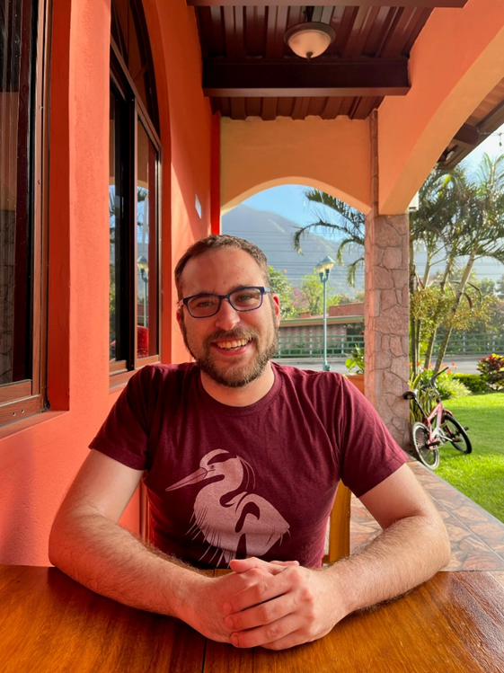 A smiling white man with brown hair, brown beard, and glasses in a red t-shirt in a tropical location